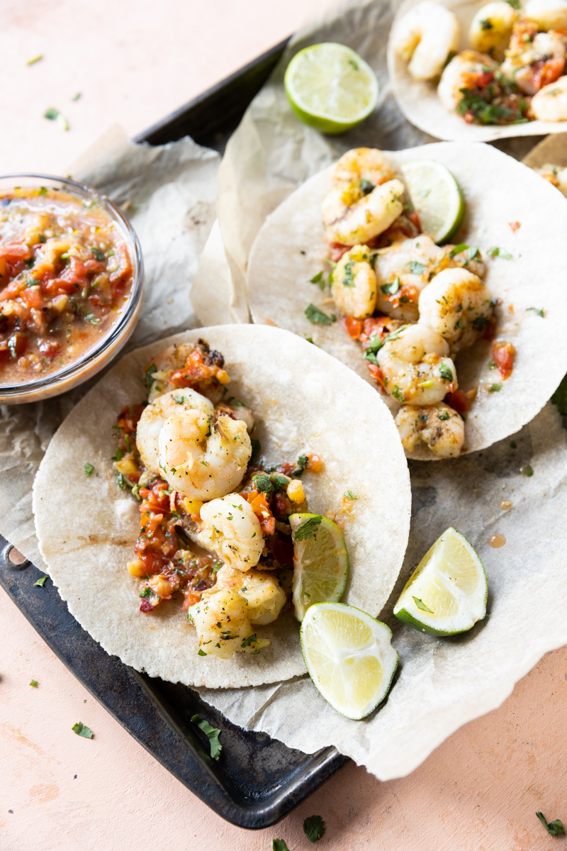 Tacos on a baking sheet with limes, salsa, and parchment paper.