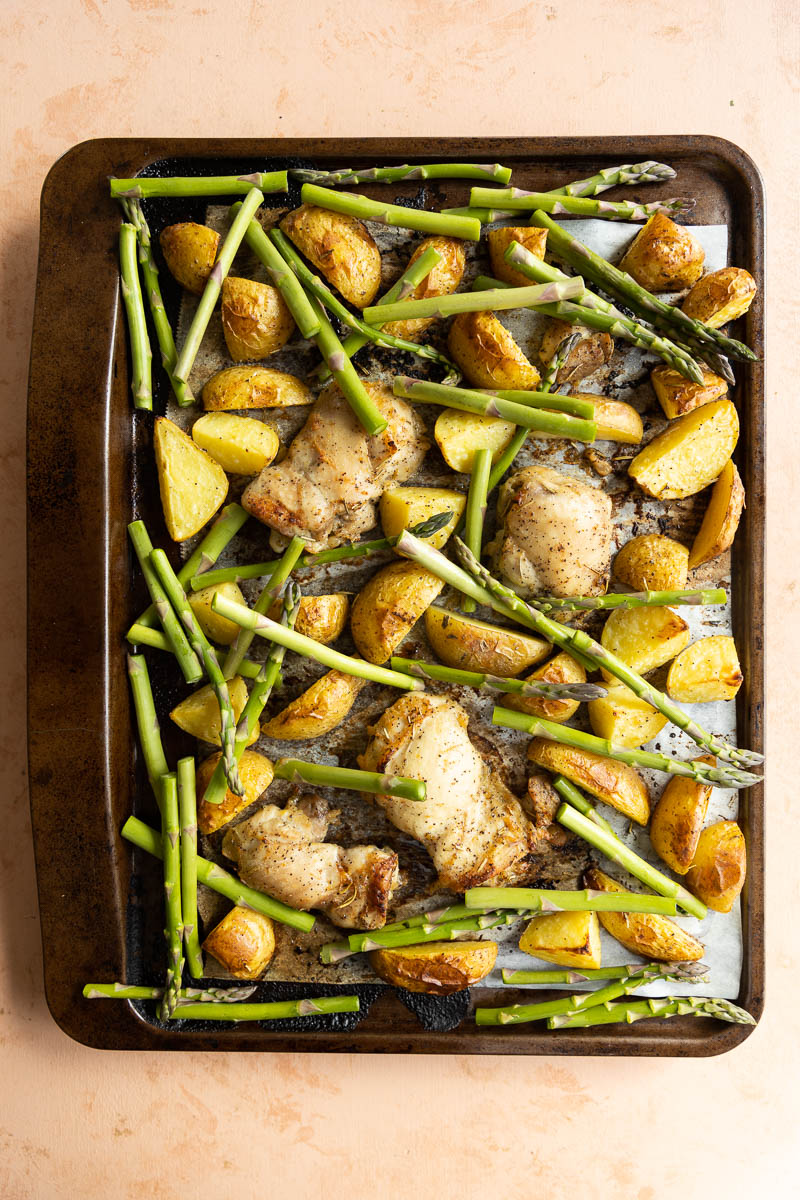 Cooked chicken and potatoes with uncooked asparagus on a parchment paper lined tray.