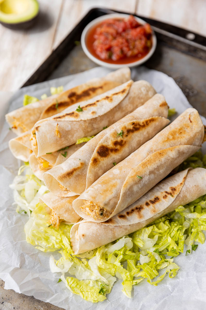 Taquitos stacked up on a lettuce bed with salsa on the side.