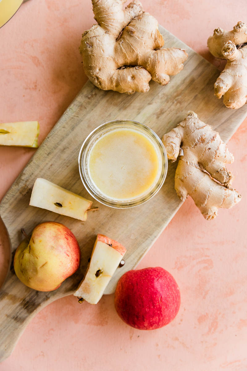 Ginger shot in a small glass surrounded by apple pieces and ginger root.
