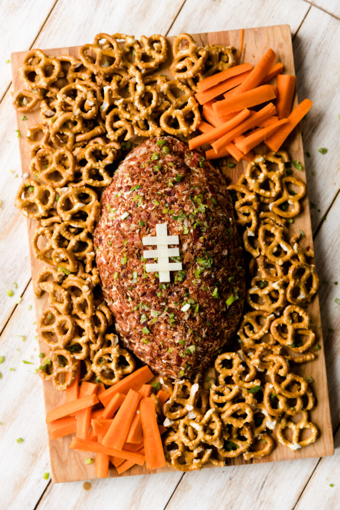 Cheese ball covered in bacon on board with pretzels and carrot sticks.