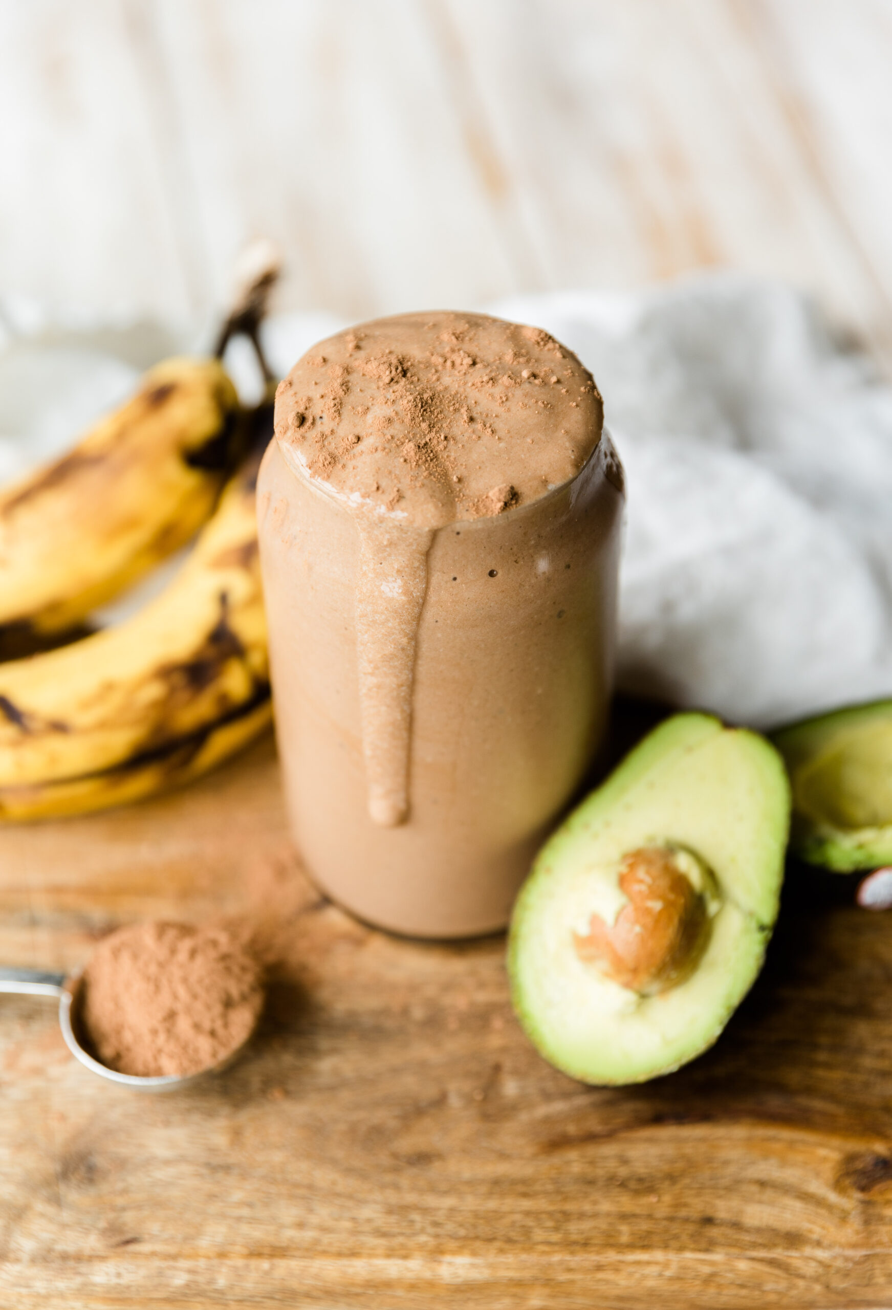 Chocolate protein shake in glass cup with half of an avocado on the side and ripe bananas.