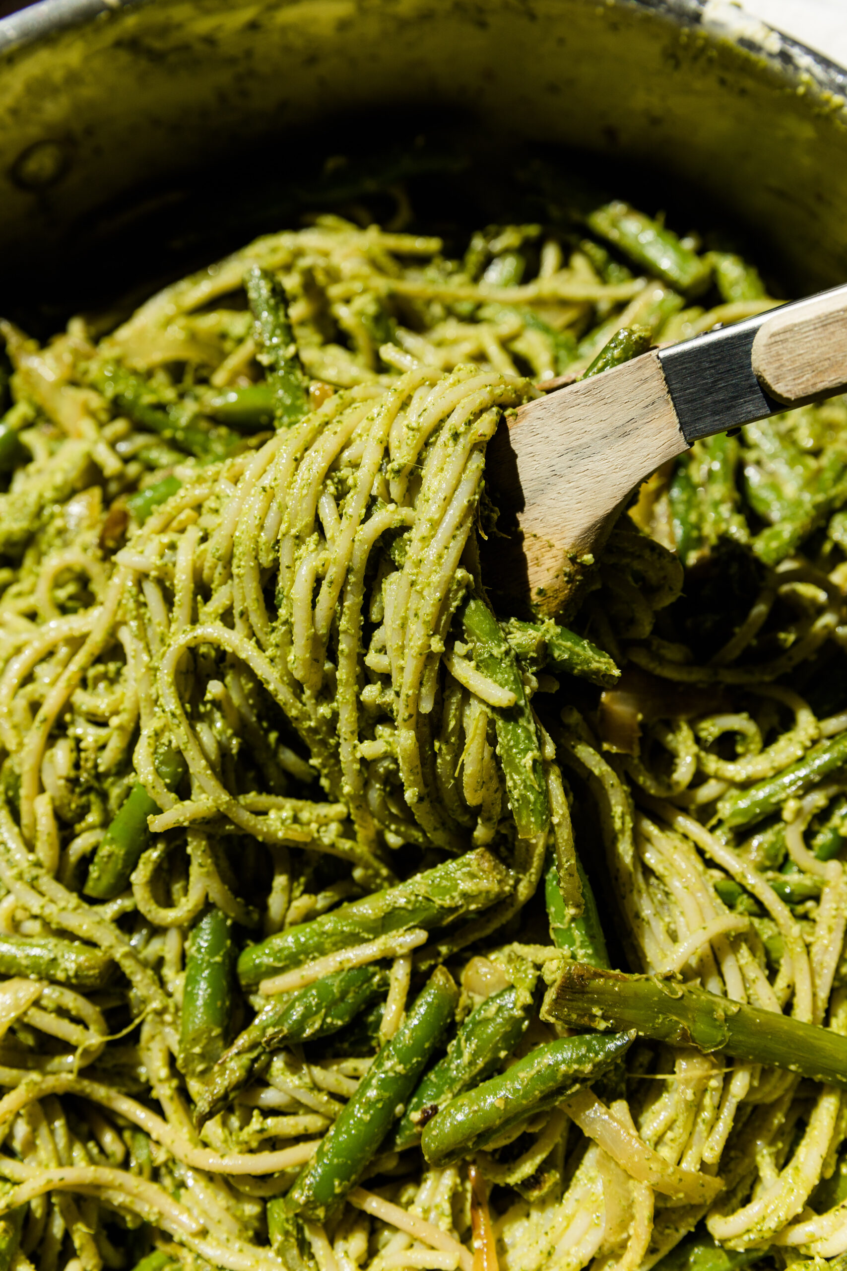 Wooden tongs grabbing spaghetti with pesto and veggies.