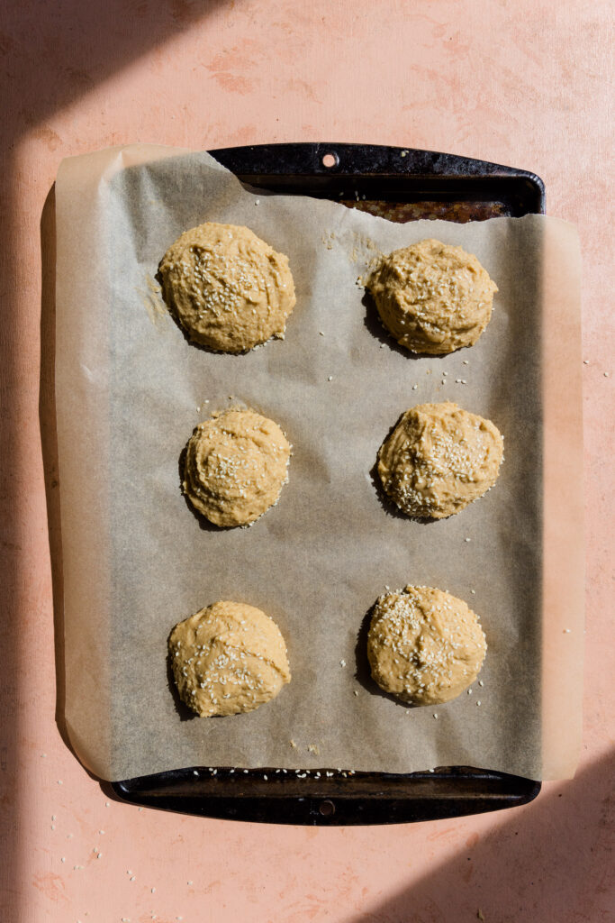 Six hamburger buns on a parchment paper lined baking sheet before going in the oven.
