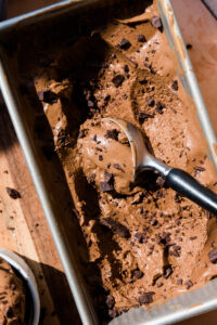 Dairy-free chocolate ice cream melting in loaf pan with ice cream scooper and chocolate chunks.
