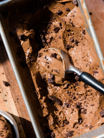 Dairy-free chocolate ice cream melting in loaf pan with ice cream scooper and chocolate chunks.
