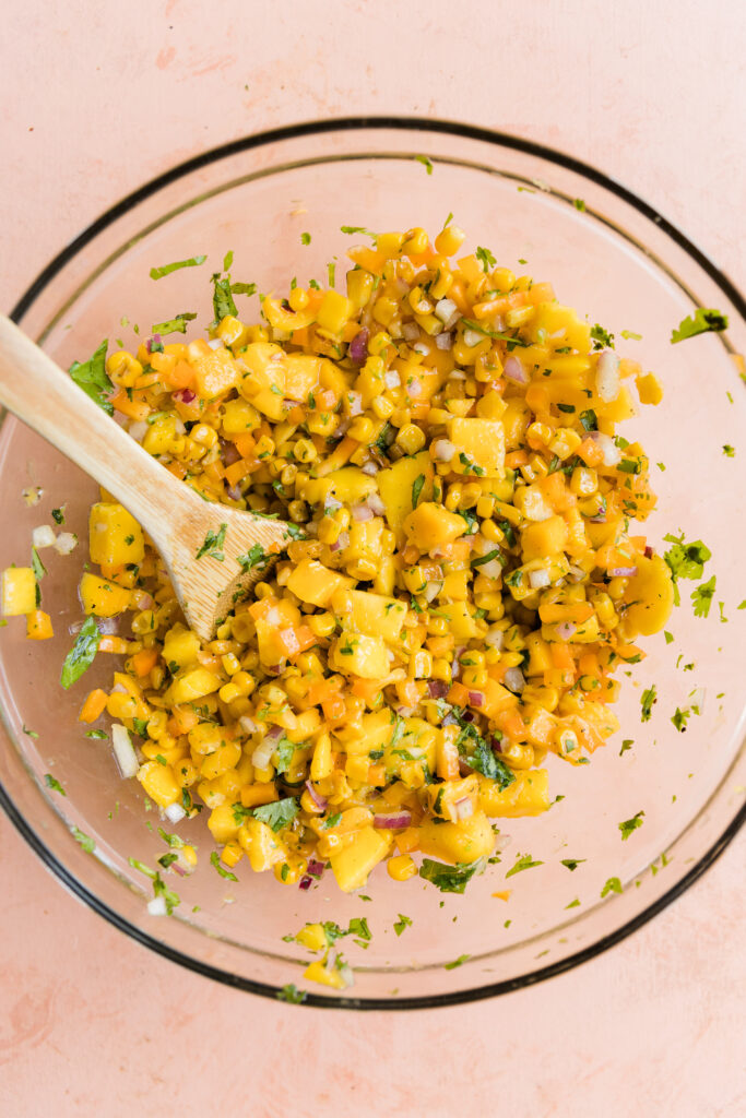 Mango corn salsa mixed up in glass bowl with wooden spoon.