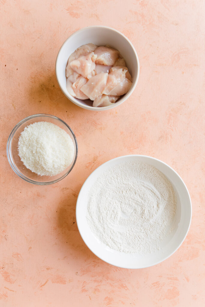 Three bowls with raw chicken pieces, shredded coconut, and cassava flour mixture.