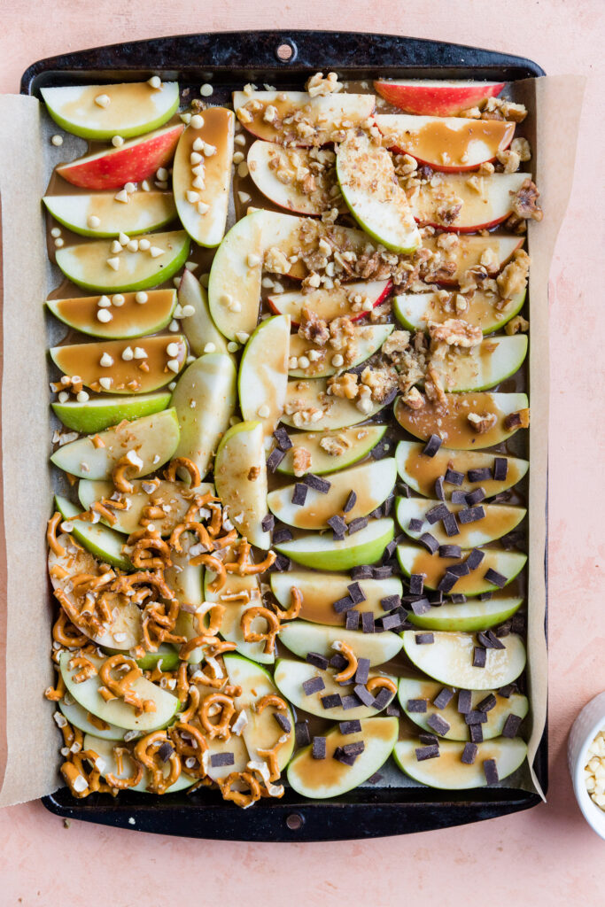 Apple slices on a sheet pan covered in dairy free caramel and gluten free pretzels, dark chocolate chips, white chocolate chips, and walnuts.